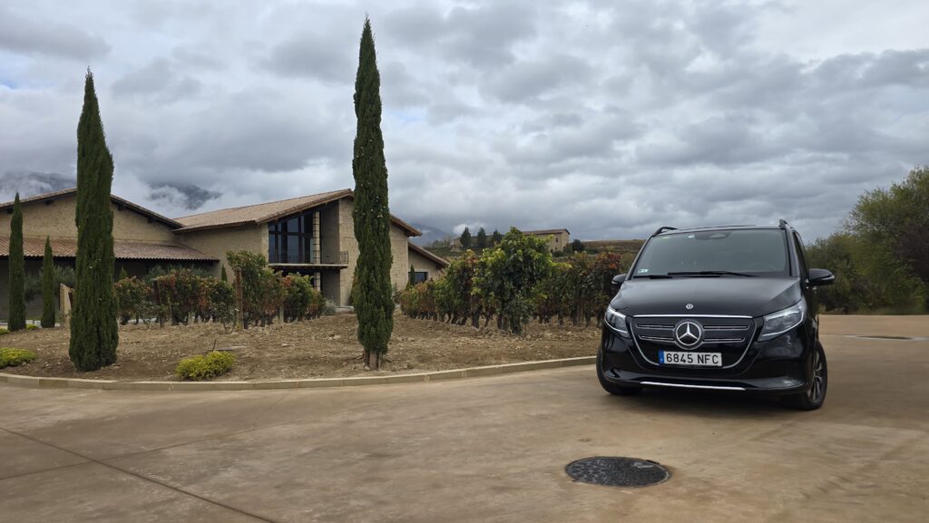 Una Mercedes-Benz Clase V negra aparcada frente a la entrada de la bodega Viñedos de Páganos, con su distintiva arquitectura de piedra integrada en el viñedo y las colinas cubiertas de vides.