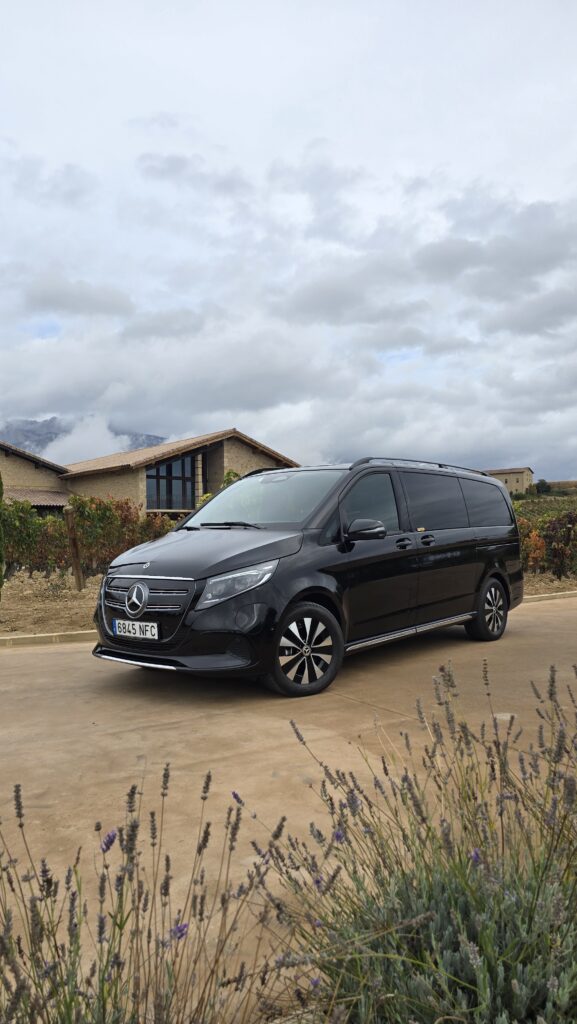 Una Mercedes-Benz clase V negra aparcada en una carretera entre viñedos, con lavanda en primer plano y una bodega tradicional al fondo.