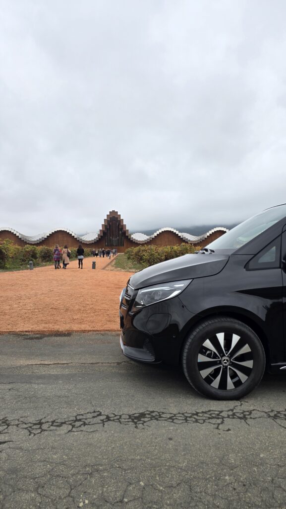 Una Mercedes-Benz Clase V negra aparcada frente a la icónica Bodega Ysios, diseñada por Calatrava, con su característico techo ondulado y una hilera de cipreses.