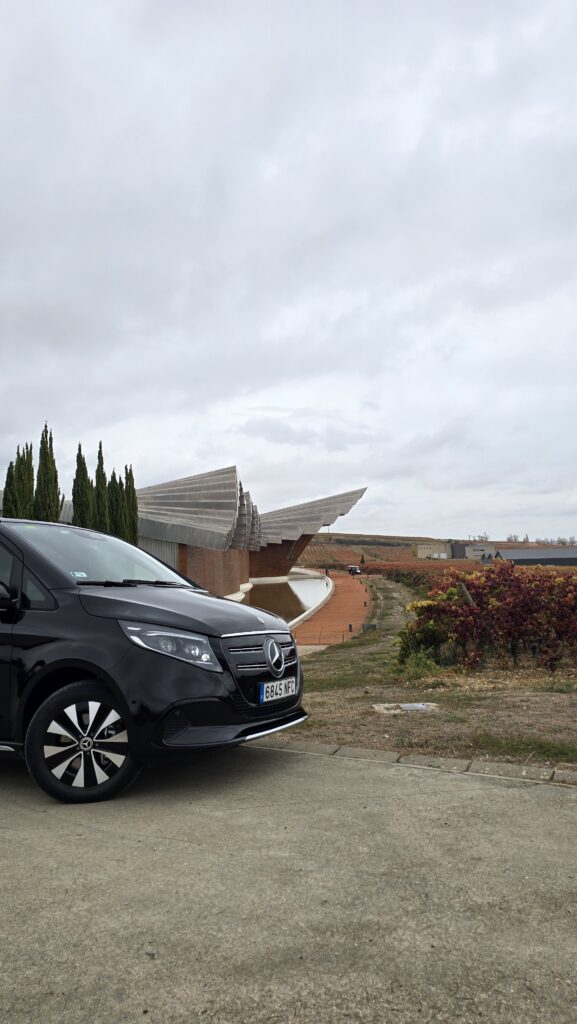 Vista frontal de cerca de una Mercedes-Benz Clase V negra, mostrando la parrilla y los faros, con un viñedo y una bodega tradicional al fondo.