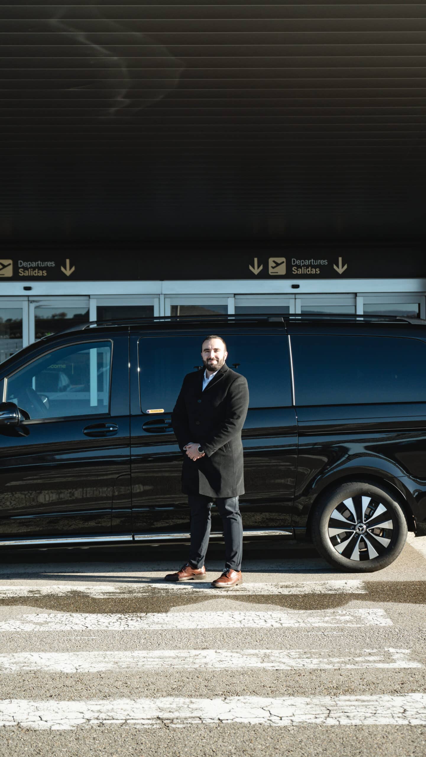 Professional bilingual private chauffeur standing next to a black luxury Mercedes V-Class at the airport in La Rioja.